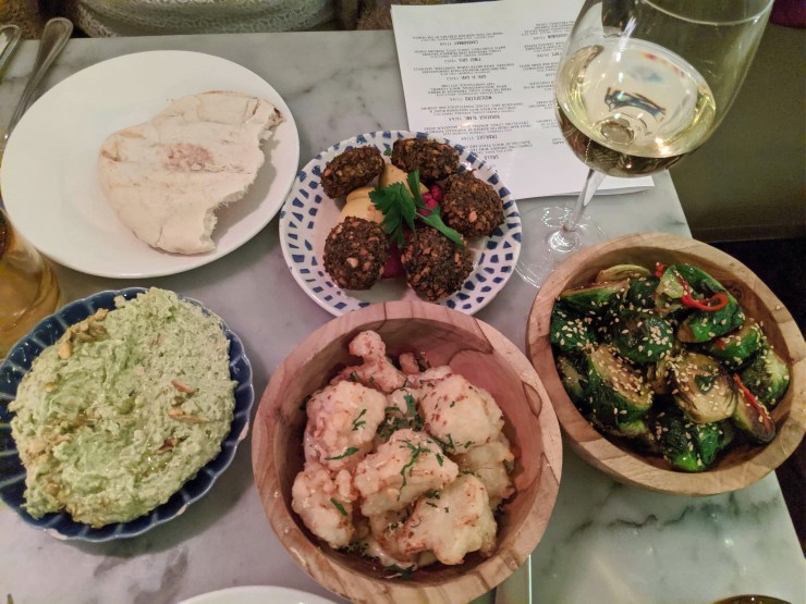 Tabletop with glass of white wine, brussels sprouts, pita bread, falafel, crispy cauliflower, and whipped feta and pistachio dip